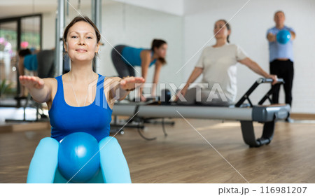 People of different ages pressing core and clutching mini Pilates balls between legs during Pilates training together 116981207