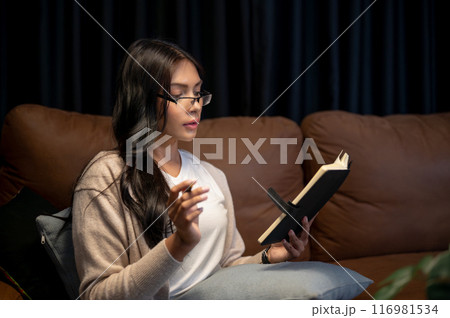 A beautiful Asian woman wearing eyeglasses is reading a book, writing in her planner on a sofa. 116981534