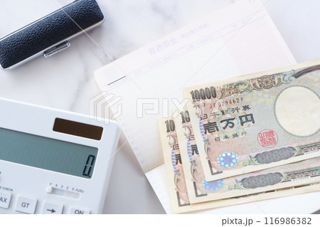 テーブルの上に開いた状態で置かれた銀行の預金通帳と、1万円札、電卓、印鑑 116986382