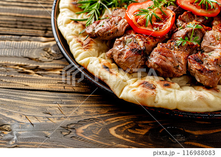 pieces of meat fried on a spit served on thick pita bread with the addition of herbs and vegetables, close-up on a dish on an old dark rough wooden table 116988083