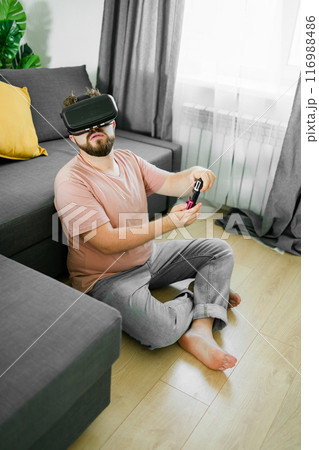 Young man spending time at home, sitting on a couch in apartment and playing arcade car video games on console. Male using controller to play street racing drift simulator. 116988486