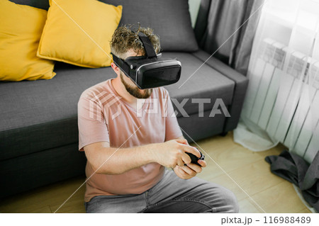 Cheerful young man seated on a sofa playing video games at home. Gaming addiction and entertainment Cheerful young man seated on a sofa playing video games at home. Gaming addiction and entertainment 116988489