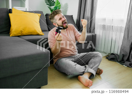 Young man spending time at home, sitting on a couch in apartment and playing arcade car video games on console. Male using controller to play street racing drift simulator. 116988494