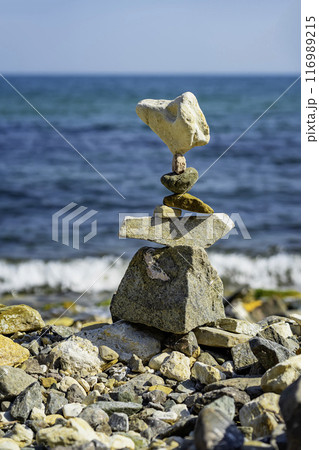 Balanced stone tower stands precariously on rocky beach, with blue ocean and foamy waves in the background Balanced stone tower stands precariously on rocky beach, with blue ocean and foamy waves in the background 116989215