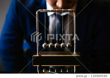 Unrecognizable businessman pulling small metal ball of Newton cradle and observing momentum over glass tabletop 116989580