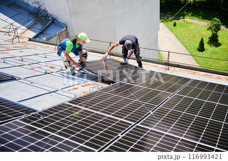 Workers building solar panel system on metal rooftop of house. Two men installers installing photovoltaic solar module outdoors. Alternative, green and renewable energy generation concept. Workers building solar panel system on metal rooftop of house. Two men installers installing photovoltaic solar module outdoors. Alternative, green and renewable energy generation concept. 116993421