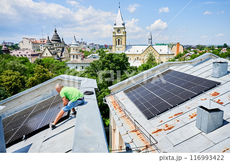 Worker building solar panel system on metal rooftop of house in urban setting. Man installer installing photovoltaic solar module outdoors. Alternative, green and renewable energy generation concept. Worker building solar panel system on metal rooftop of house in urban setting. Man installer installing photovoltaic solar module outdoors. Alternative, green and renewable energy generation concept. 116993422