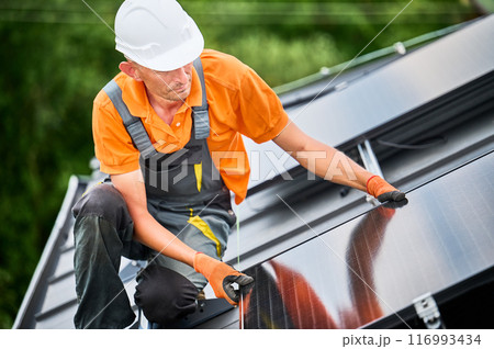 Worker building solar panel system on rooftop of house. Man engineer in helmet installing photovoltaic solar module outdoors. Alternative, green and renewable energy generation concept. Worker building solar panel system on rooftop of house. Man engineer in helmet installing photovoltaic solar module outdoors. Alternative, green and renewable energy generation concept. 116993434