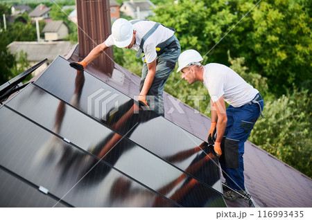 Workers building solar panel system on rooftop of house. Two men installers in helmets installing photovoltaic solar module outdoors. Alternative, green and renewable energy generation concept. Workers building solar panel system on rooftop of house. Two men installers in helmets installing photovoltaic solar module outdoors. Alternative, green and renewable energy generation concept. 116993435