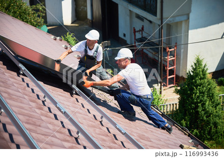 Workers building solar panel system on rooftop of house. Two men installers in helmets installing photovoltaic solar module outdoors. Alternative, green and renewable energy generation concept. 116993436