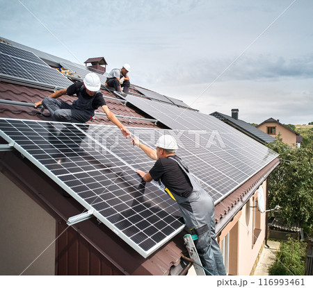 Men technicians mounting photovoltaic solar moduls on roof of house. Engineers in helmet installing solar panel system outdoors. Concept of alternative and renewable energy. Aerial view. Men technicians mounting photovoltaic solar moduls on roof of house. Engineers in helmet installing solar panel system outdoors. Concept of alternative and renewable energy. Aerial view. 116993461
