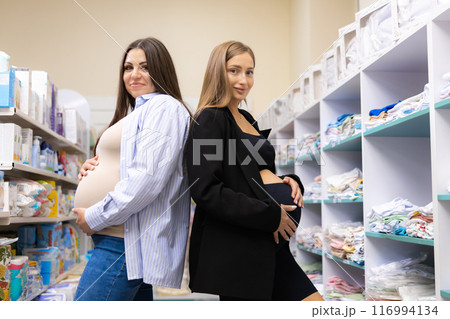 Two pregnant friends shopping together in clothes store. Mothers doing shopping in baby shop. Baby clothes sale and pregnancy concept. 116994134