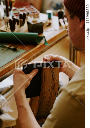 Over shoulder shot of man sitting at table and sewing stitches on leather Over shoulder shot of man sitting at table and sewing stitches on leather 116996880
