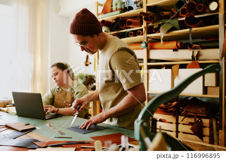 Male tailor preparing leather workpieces while his young female intern using laptop in background Male tailor preparing leather workpieces while his young female intern using laptop in background 116996895