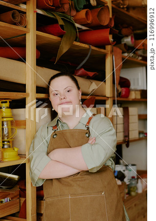 Talented female tailor with down syndrome dressed in t-shirt and chamois apron leaning on wooden shelves in atelier 116996912