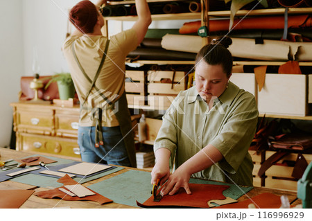 Female tailor with down syndrome cutting out leather workpieces while male tailor looking for material on wooden shelves Female tailor with down syndrome cutting out leather workpieces while male tailor looking for material on wooden shelves 116996929