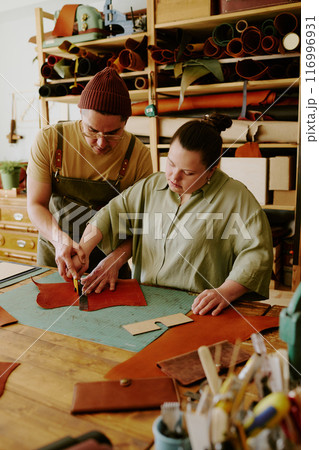 Highly skilled male tailor helping his new female intern with down syndrome to cut out leather workpiece Highly skilled male tailor helping his new female intern with down syndrome to cut out leather workpiece 116996931