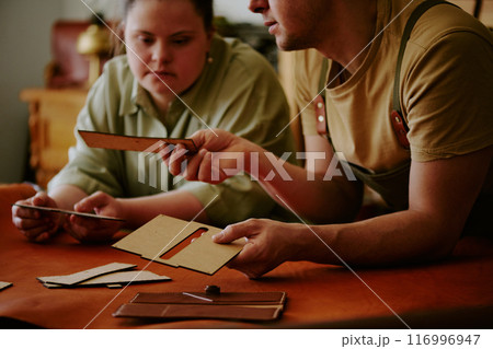 Male tailor demonstrating wooden rectangular shape to his female coworker with down syndrome while they leaning on wooden table covered with leather 116996947
