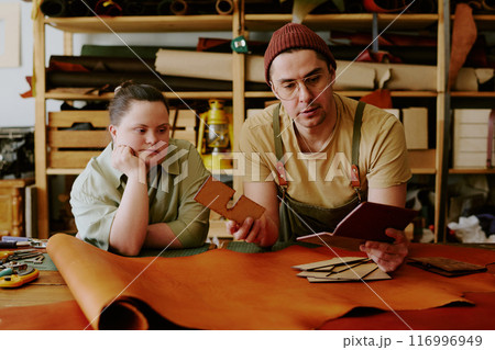 Caucasian male tailor and female client with down syndrome comparing leather workpieces with tailors notes Caucasian male tailor and female client with down syndrome comparing leather workpieces with tailors notes 116996949