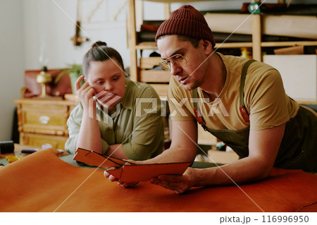 Tailor and client with down syndrome leaning on old table while examining closely leather workpieces 116996950