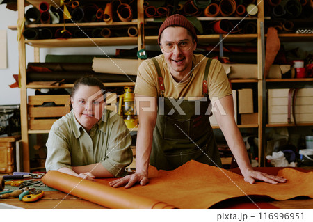 Portrait of smiling male tailor and his female client with down syndrome leaning on wooden table covered with leather workpieces while in atelier Portrait of smiling male tailor and his female client with down syndrome leaning on wooden table covered with leather workpieces while in atelier 116996951