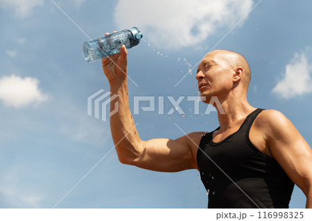 Sweaty handsome sporty man refreshing himself water from bottle against blue sky at hot summer after training outdoors. Concept water balance. Close up. Banner. 116998325