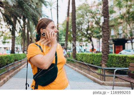 A portrait of a smiling beautiful woman talking on the phone in the public park. 30s cheerful woman tourist dressed in casual look resting outdoors A portrait of a smiling beautiful woman talking on the phone in the public park. 30s cheerful woman tourist dressed in casual look resting outdoors 116999203