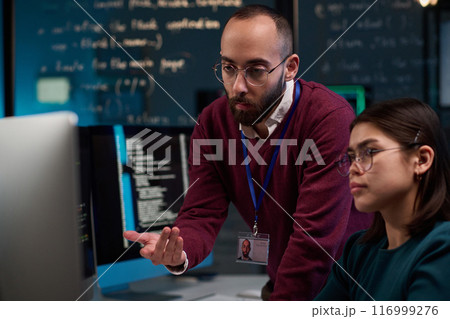 Portrait of bearded IT developer pointing at computer screen while advising young trainee in office with code lines in background Portrait of bearded IT developer pointing at computer screen while advising young trainee in office with code lines in background 116999276