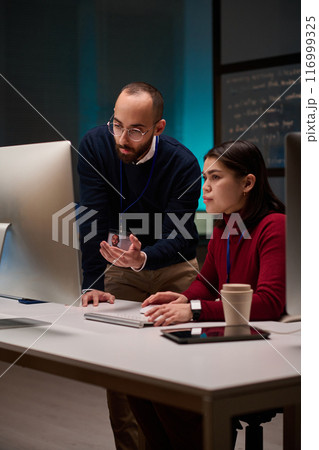 Vertical portrait of cybersecurity team of two people using computer together in office lit by blue light 116999325