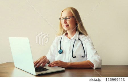 Happy woman doctor working with laptop computer while sitting at desk in medical clinic office 116999428