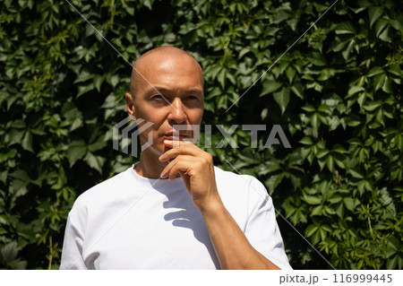 Outdoor portrait of thinking and looking away caucasian adult man wearing white t-shirt against plant wall of green leaves. Eco concept. Sustainable lifestyle. 116999445