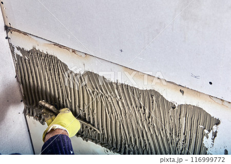 Builder tiler applies adhesive cement mortar to wall using comb trowel. Builder tiler applies adhesive cement mortar to wall using comb trowel. 116999772