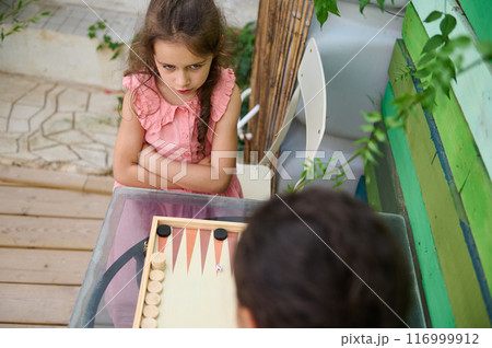 Young girl playing backgammon with friend outdoors, expression of concentration Young girl playing backgammon with friend outdoors, expression of concentration 116999912
