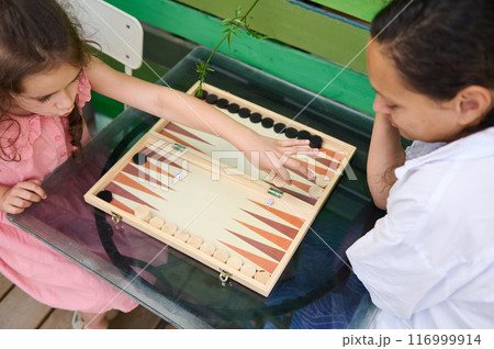 Mother and daughter playing an engaging game of backgammon on a glass table outdoors 116999914