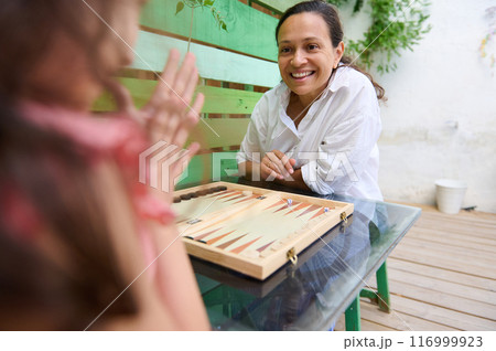 Mother and daughter playing board game on outdoor patio table and enjoying quality time 116999923