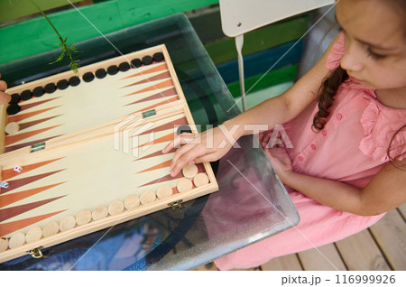 Young girl playing backgammon outdoors on a glass table wearing a pink dress 116999926