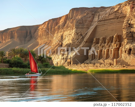 A sailboat with a red sail glides on a river beside a cliff with large ancient Egyptian temple carvings. 117002352