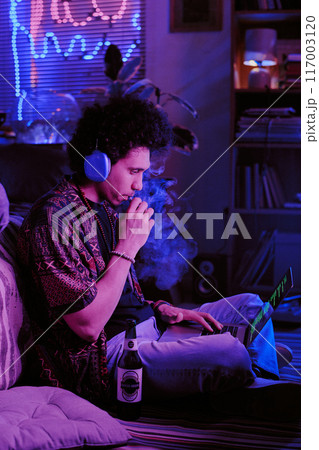 Curly-haired developer taking inhalation of sweet vape while checking codes on laptop 117003120