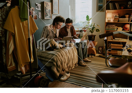 University Students sitting on bed and comparing different databases while working on their startup together in messy dorm room 117003155