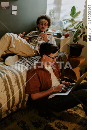 Best friends resting after lectures in dorm room, curly-haired student playing guitar while his friend playing computer games Best friends resting after lectures in dorm room, curly-haired student playing guitar while his friend playing computer games 117003162