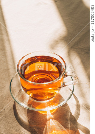 A cup of tea in glass cup and saucer illuminated by warm sunlight on a light table with a simple tablecloth. Vertical frame, copy space A cup of tea in glass cup and saucer illuminated by warm sunlight on a light table with a simple tablecloth. Vertical frame, copy space 117003405