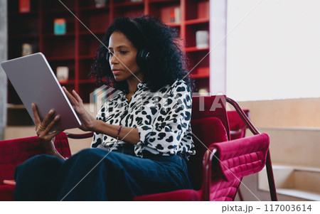 Adult Black female in casual attire, focused on tablet, with headphones, indoors, concept of digital learning 117003614