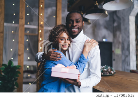 joyful Black couple embraces in cozy indoor , husband man presenting pink gift box. 117003618
