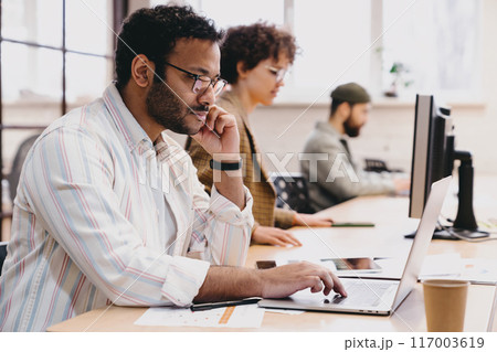 Smiling ethnic man working on laptop with colleagues in office 117003619
