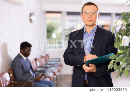 Adult man in glasses with documents stands in reception 117005950