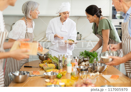 Serious young woman chef of cooking course teaching attendees how to cook salmon Serious young woman chef of cooking course teaching attendees how to cook salmon 117005970
