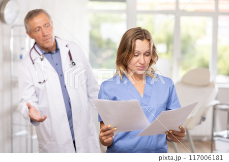 Upset female doctor with papers stading in office with disgruntled chief 117006181