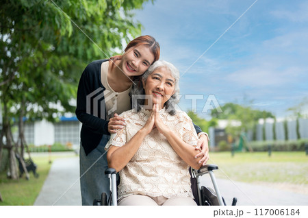 A lovely granddaughter is hugging her grandma, who is in a wheelchair, while walking her in a park. 117006184