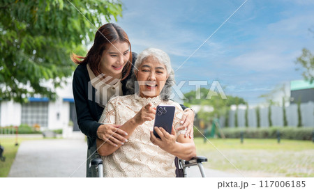 A lovely Asian granddaughter is hugging her grandma who is using a smartphone in a wheelchair. 117006185