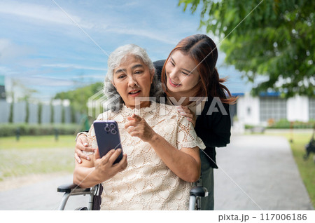 A lovely Asian granddaughter is hugging her grandma who is using a smartphone in a wheelchair. 117006186